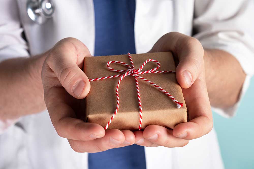 Arzt hält Weihnachts-Geschenk auf hellblauem Hintergrund